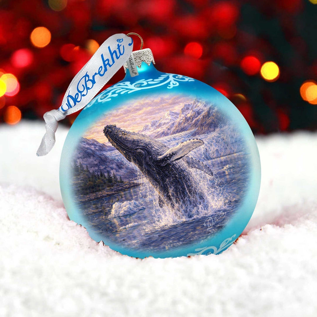 A blue ornament with a painting of a whale leaping out of the water, set against a snowy background with red and orange lights in the background.