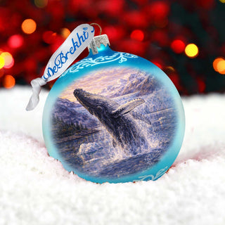A blue ornament with a painting of a whale leaping out of the water, set against a snowy background with red and orange lights in the background.