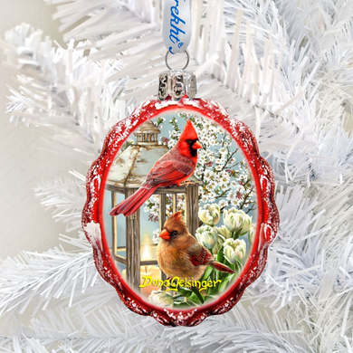 A red ornament with a picture of two red cardinals on a snowy branch, hanging on a white tree branch.
