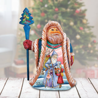 A colorful figurine of Santa Claus holding a reindeer and a blue staff, set against a blurred Christmas tree background.