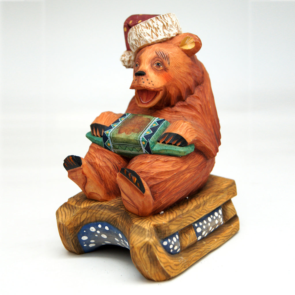A wooden bear figurine wearing a hat and holding a musical instrument, sitting on a wooden sled.