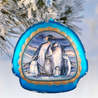 A blue ornament with a picture of a group of penguins on it, hanging on a tree branch covered in snow.