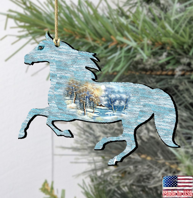 A wooden ornament in the shape of a horse, with a winter landscape scene inside, hanging on a tree branch.