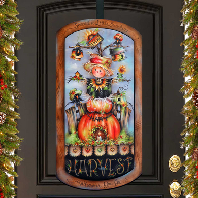 a decorative sign hanging on a door, depicting a scarecrow with a pumpkin and other fall-themed elements.
