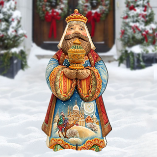 A colorful, hand-painted wooden statue of a king or ruler, holding a golden object, stands in the snow in front of a building with wreaths and Christmas decorations.