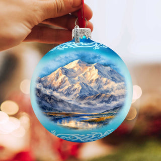 A hand holding a blue ornament with a painting of a mountain landscape.