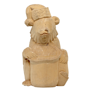 A wooden carving of a bear wearing a hat and holding a hat in its paws, sitting on a hat.