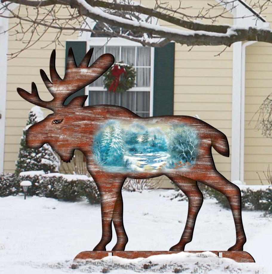 A wooden moose sculpture stands in a snowy landscape, with a wreath hanging on a window behind it.