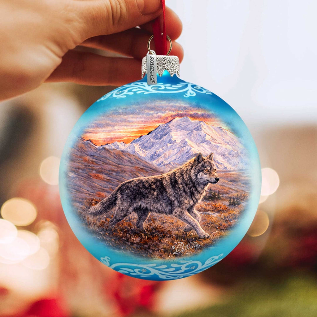 A hand is holding a blue ornament with a painting of a wolf in a snowy mountain landscape.