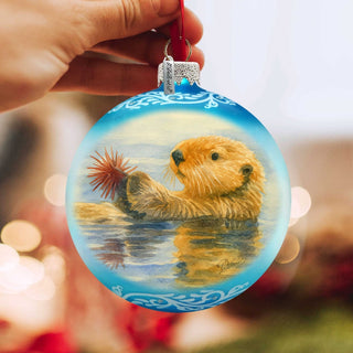 A hand holding a blue ornament with a painting of a sea otter in the water.