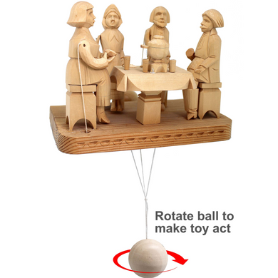 A wooden toy with four figures, including a woman, a man, a child, and a dog, sitting around a table with a bowl of food. The toy is suspended from a string, and there is a text overlay instructing to rotate the ball to make the toy act.