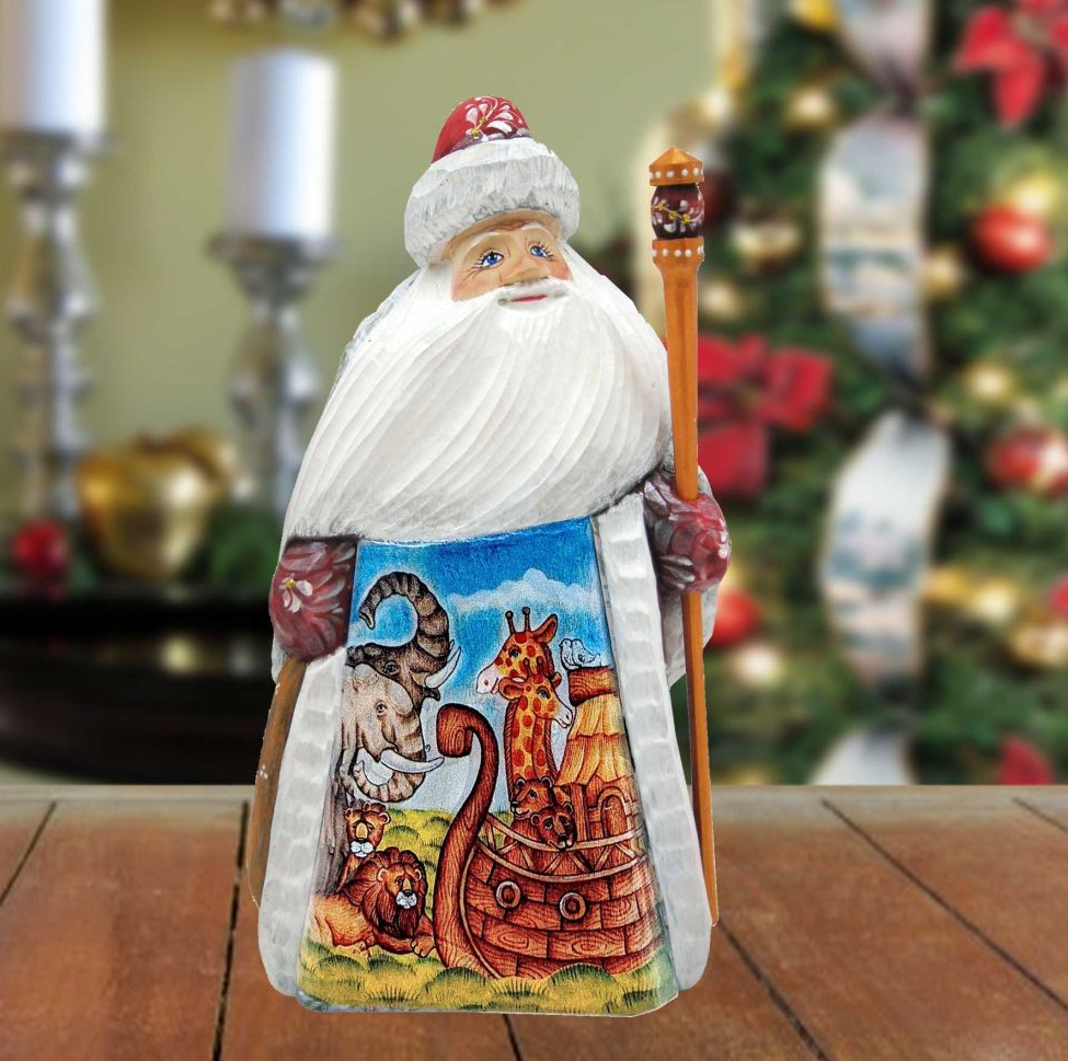 A wooden Santa Claus figurine holding a staff and a decorated nativity scene, with a blurred Christmas tree in the background.