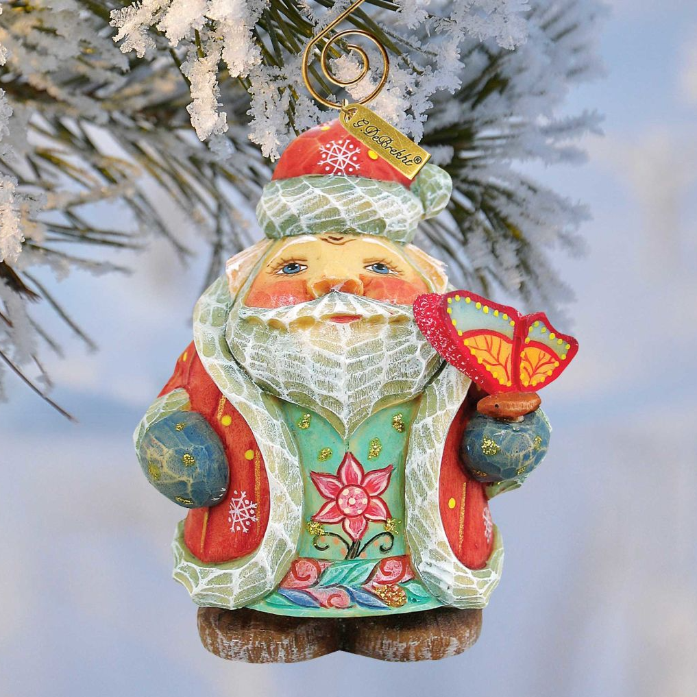 A colorful ornament of a smiling Santa Claus figure holding a butterfly, hanging from a tree branch covered in snow.