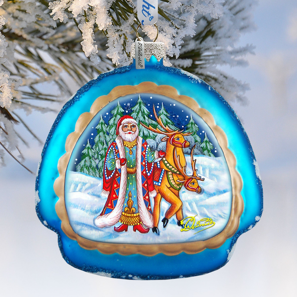 A blue ornament with a painted scene of Santa Claus and a reindeer on a snowy landscape, hanging on a tree branch.