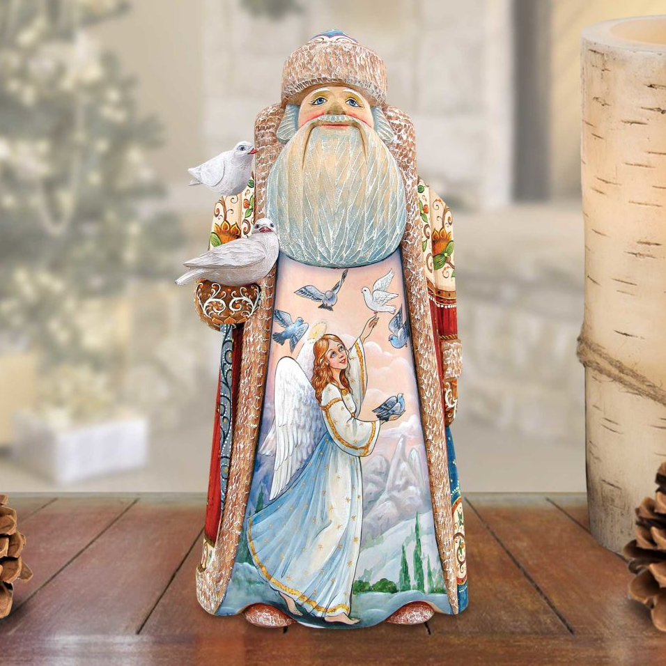 A detailed figurine of Santa Claus, adorned with a blue and white robe, holding a bird in his hand, and an angel figure, is displayed on a wooden surface.