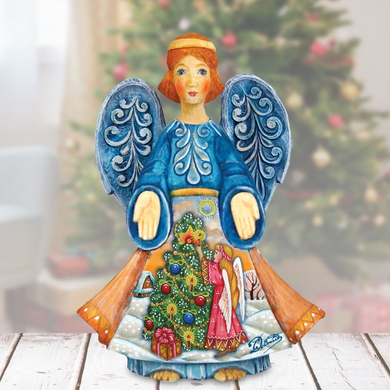 A hand-painted Christmas angel figurine with blue wings and a red head stands on a wooden surface, surrounded by a blurred Christmas tree and other holiday decorations.