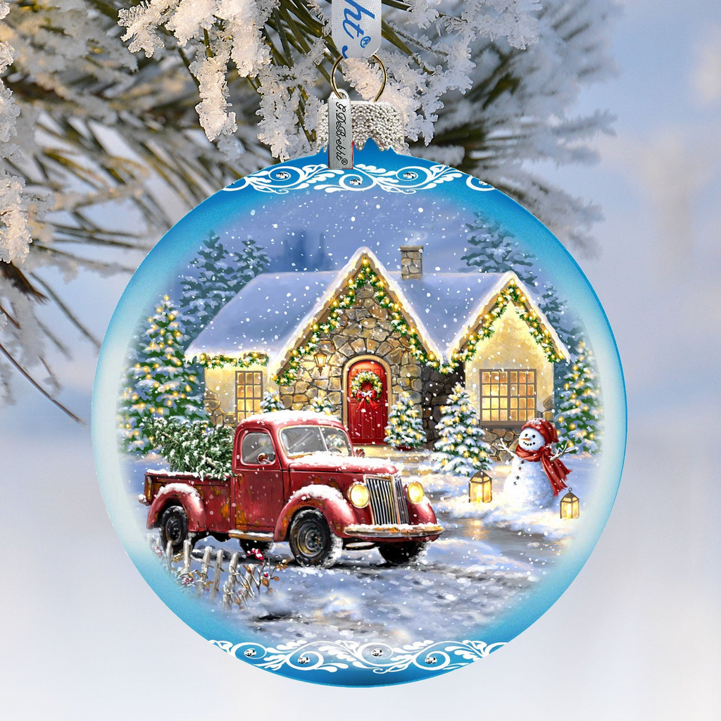 A Christmas ornament depicting a red pickup truck parked in front of a snow-covered house, with a snowman and Christmas lights visible.