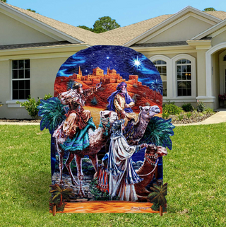 A large, colorful, and detailed cardboard cutout depicting a scene from the Christmas story of the Three Wise Men and the baby Jesus, with the figures riding on camels and the baby Jesus being carried by the wise men. The cutout is set against a backdrop of a house and a starry night sky.
