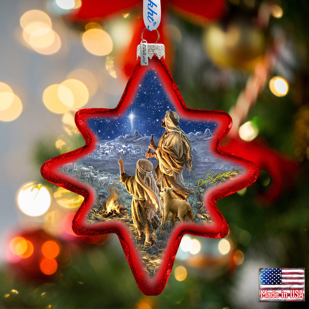 A star-shaped ornament with a painting of a shepherd and his lamb, set against a night sky backdrop, is hanging on a Christmas tree.