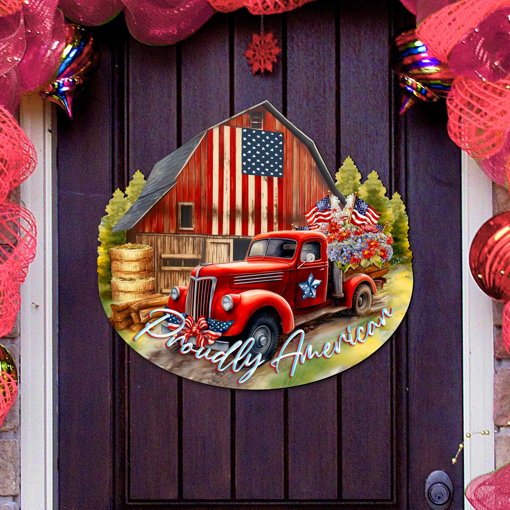 A red vintage truck is parked in front of a red barn with an American flag displayed, surrounded by a festive wreath and ornaments.
