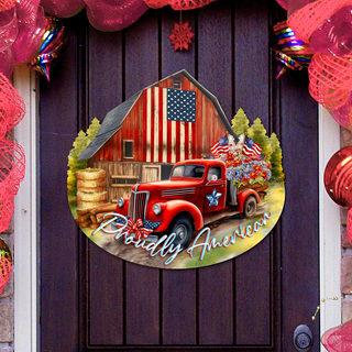 A red vintage truck is parked in front of a red barn with an American flag displayed, surrounded by a festive wreath and ornaments.
