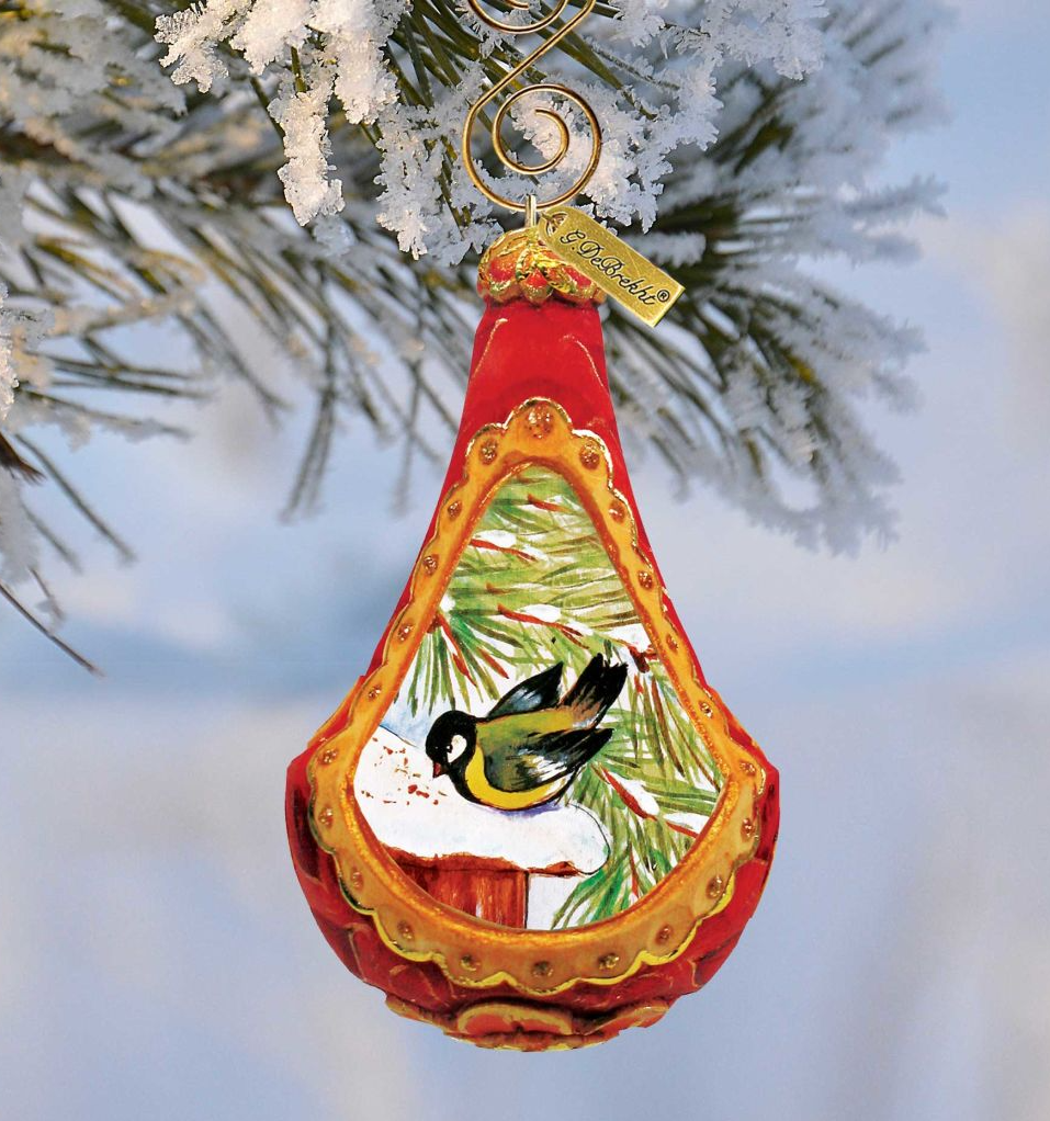 A red ornament with a gold frame hangs from a tree branch, featuring a painting of a bird perched on a branch.