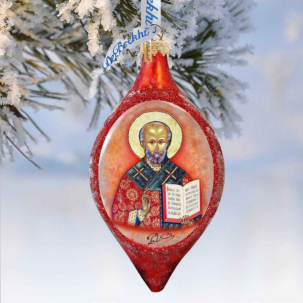 A red Christmas ornament with a depiction of a saint, hanging on a tree branch with snow on it.