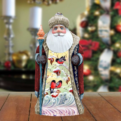 A wooden Santa Claus figurine holding a staff and wearing a colorful, patterned coat stands on a wooden surface, with a blurred Christmas tree and decorations in the background.