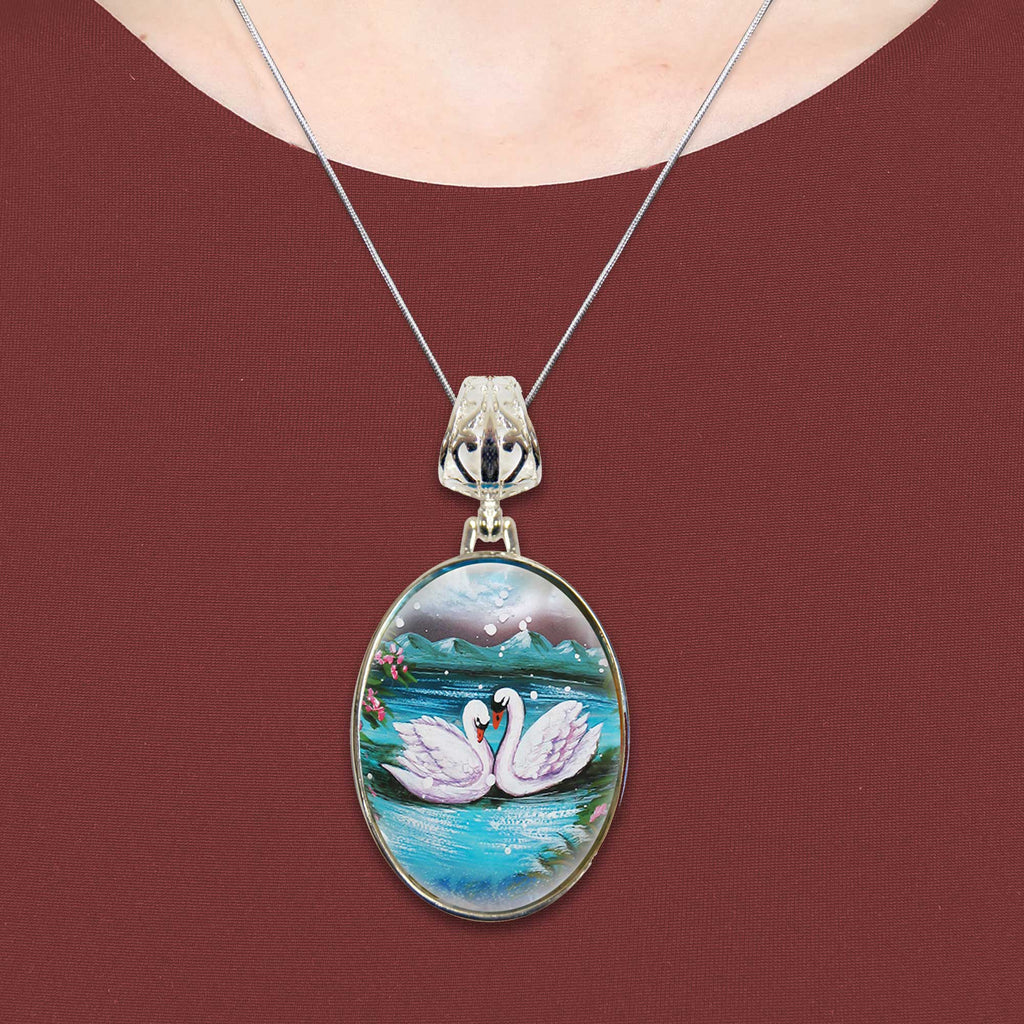 A necklace with a pendant featuring a painting of two swans on a lake.