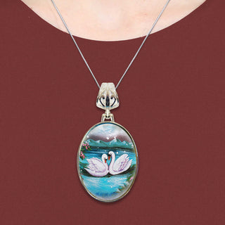 A necklace with a pendant featuring a painting of two swans on a lake.
