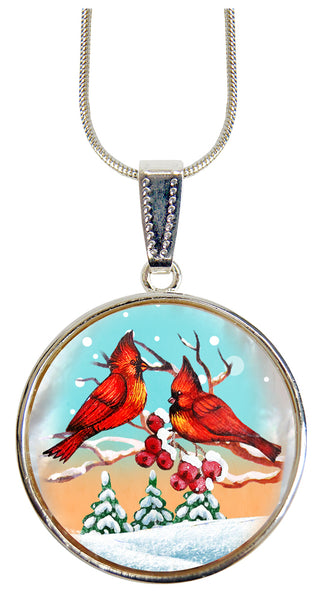 A silver pendant necklace with a circular design featuring two red cardinals perched on branches with berries, set against a snowy winter landscape.