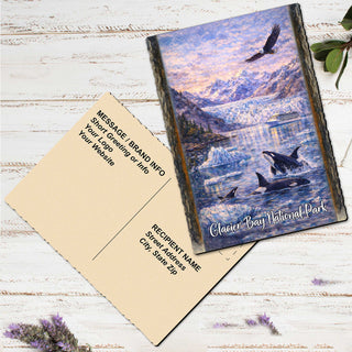 A postcard featuring an image of a scenic landscape with mountains, water, and wildlife, including an orca whale, is placed on a wooden surface alongside a blank postcard.