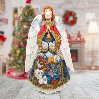 A detailed and colorful angel figurine stands in front of a Christmas tree, with a nativity scene depicted on its body.