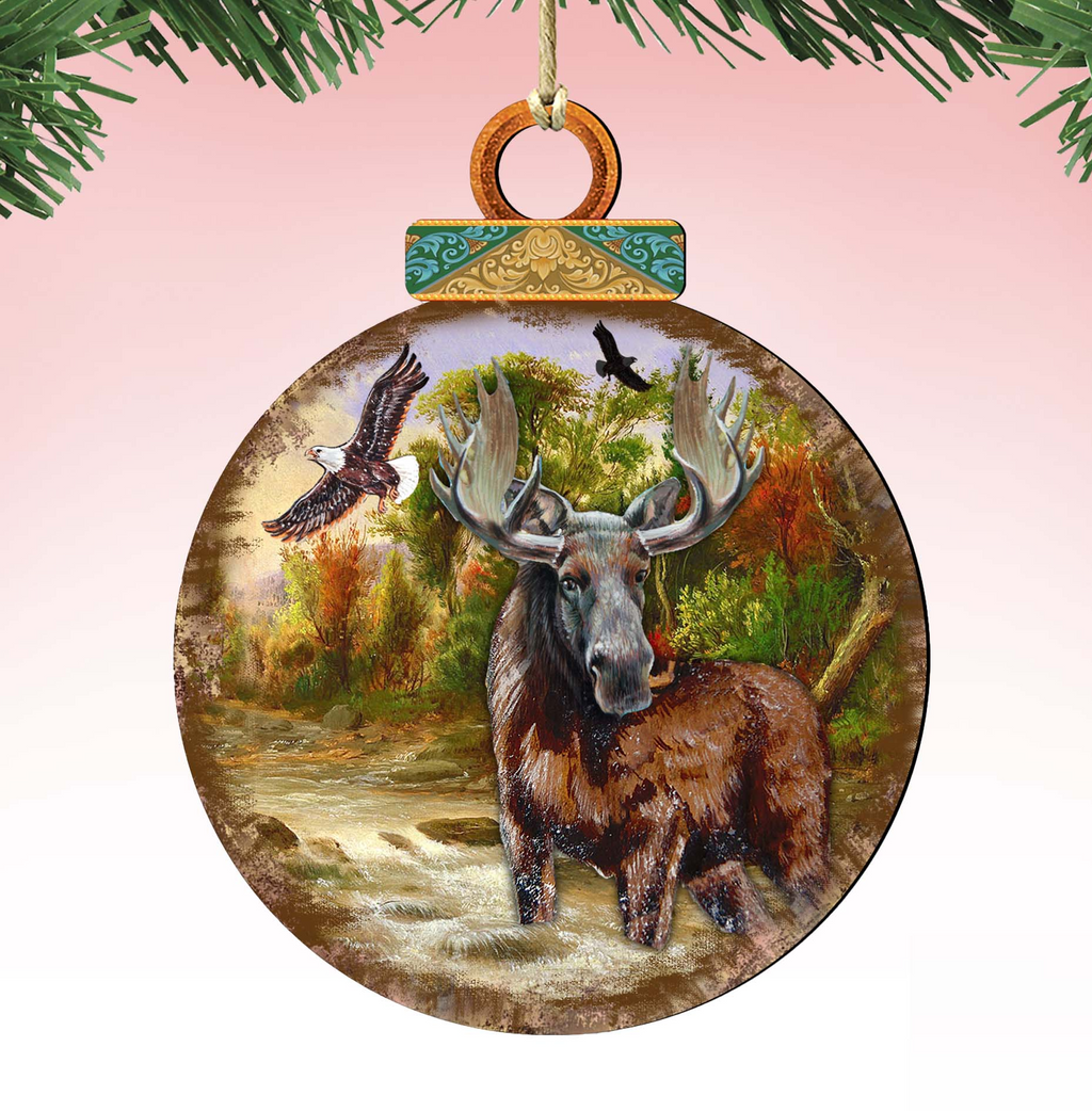 A wooden ornament featuring a moose standing in a forest, with a bird flying overhead and a stream running through the scene.