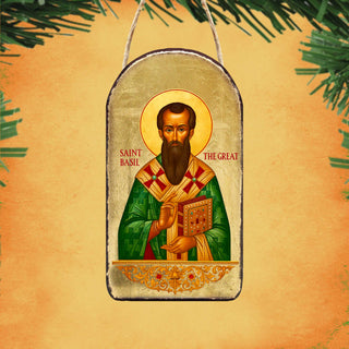 a religious icon of Saint Basil, a prominent figure in Orthodox Christianity. The icon depicts Saint Basil holding a book and wearing a green robe with a red and gold pattern. The background of the image is a warm orange color, and there are green pine branches visible in the top left corner.