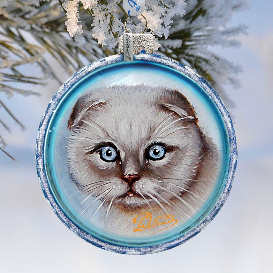 A blue ornament with a picture of a cat's face on it, hanging on a snowy branch.