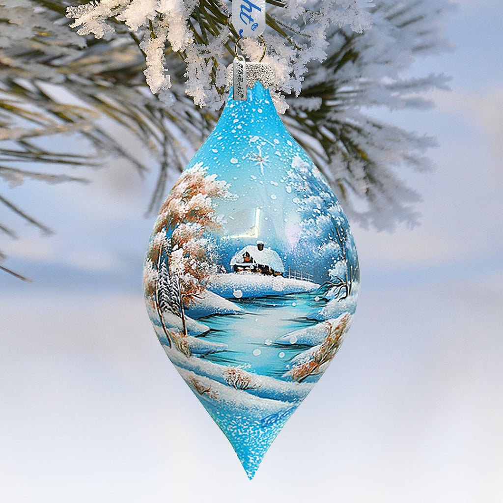 A blue Christmas ornament with a snowy landscape scene hangs from a tree branch.