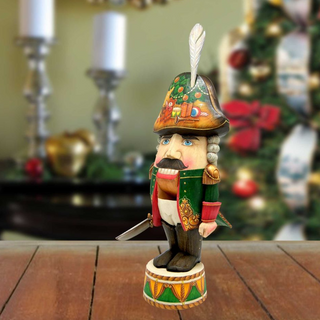 A wooden nutcracker figurine with a colorful hat and a sword stands on a wooden surface, with a blurred background featuring a Christmas tree and other holiday decorations.