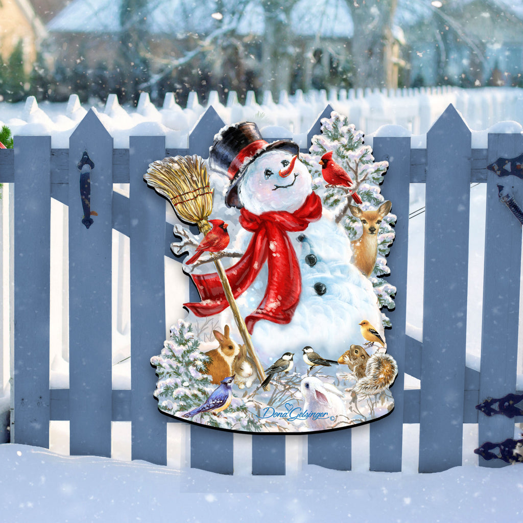 A snowman decoration with a red scarf, black top hat, and various animals, including a bird, is displayed on a blue picket fence in a snowy landscape.