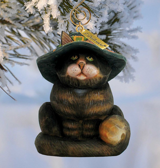 A wooden ornament in the shape of a cat wearing a green hat with a gold tag, hanging from a tree branch.