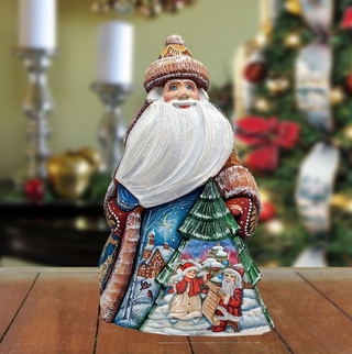 A wooden Santa Claus figurine with a white beard and hat, standing on a wooden surface. The figurine is decorated with a snowy landscape and Christmas trees, creating a festive holiday scene.