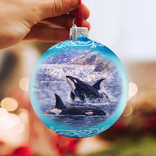A hand is holding a blue ornament with a painting of two orca whales swimming in the ocean.