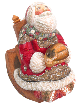 A wooden statue of Santa Claus sitting on a rocking chair, holding a small brown cat in his lap.