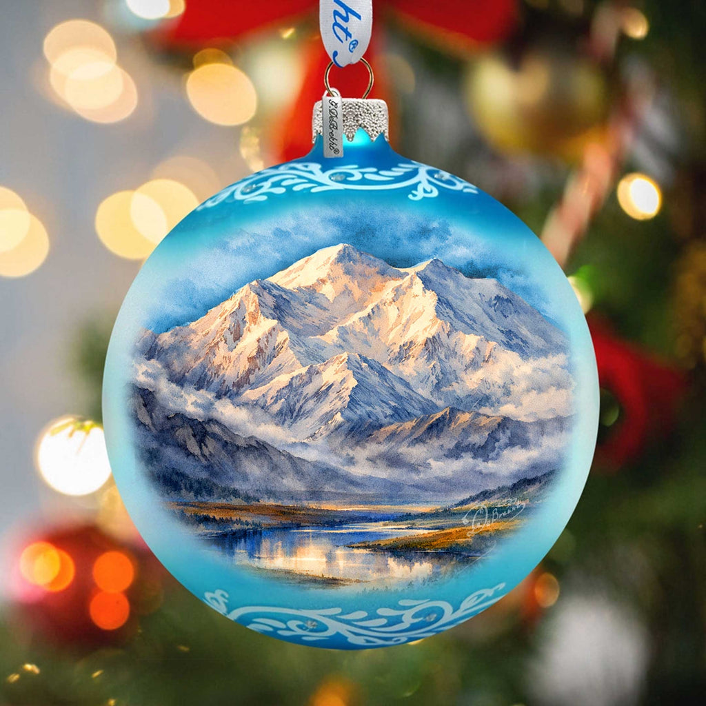 A blue Christmas ornament with a painting of a mountain landscape inside, hanging on a tree with blurred lights in the background.
