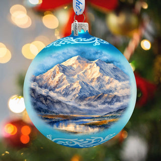 A blue Christmas ornament with a painting of a mountain landscape inside, hanging on a tree with blurred lights in the background.