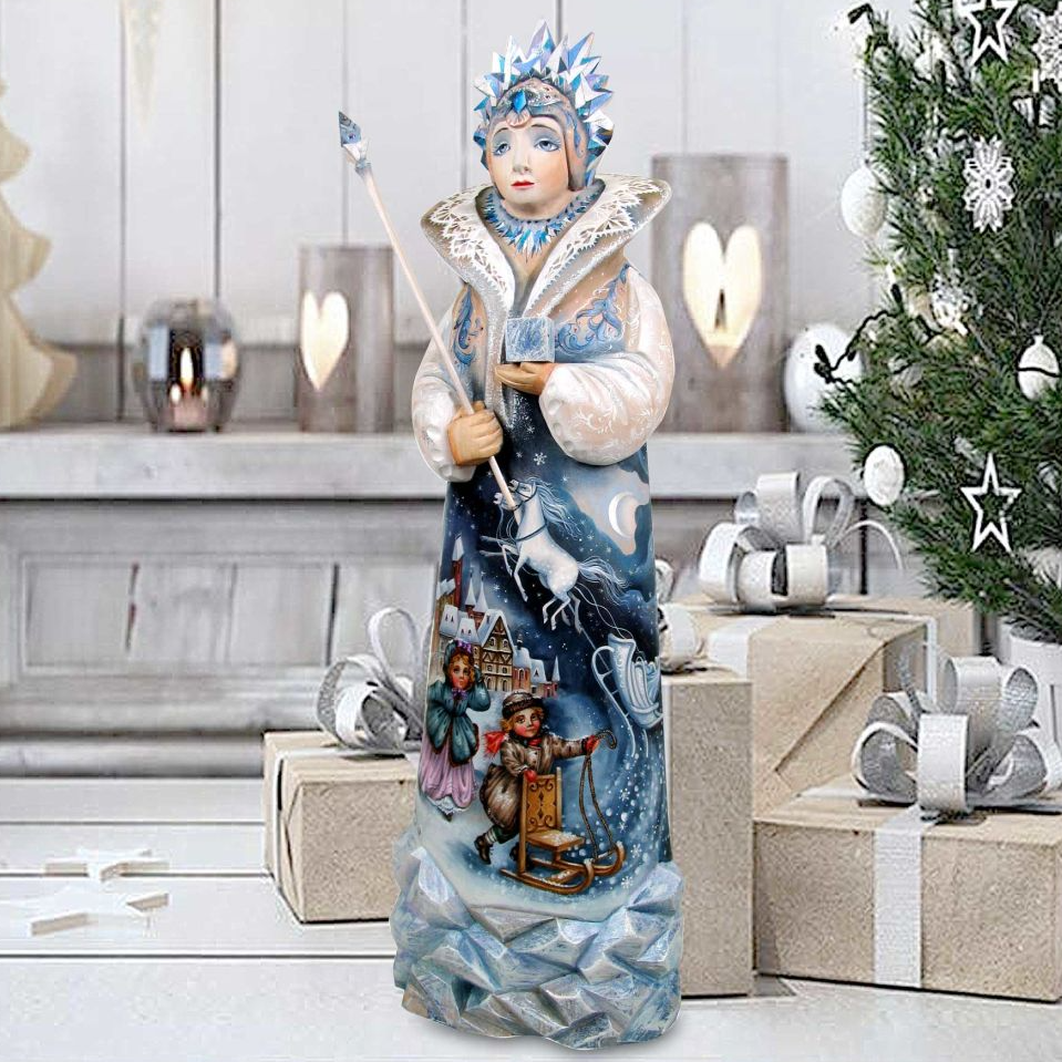 A figurine of a woman dressed in a blue and white outfit, holding a staff, stands in front of a Christmas tree and presents.