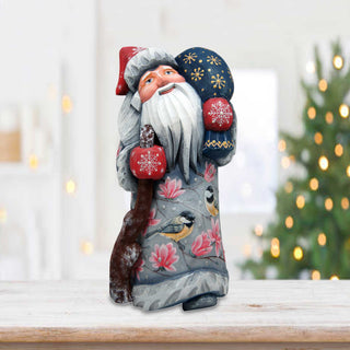 A wooden Santa Claus figurine with a blue hat and red mittens is sitting on a wooden surface, with a blurred Christmas tree in the background.