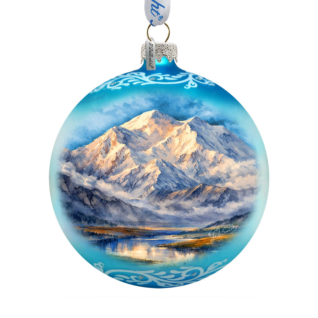 A blue glass ornament with a painting of a mountain landscape, featuring a snowy mountain peak, a lake, and a cloudy sky.