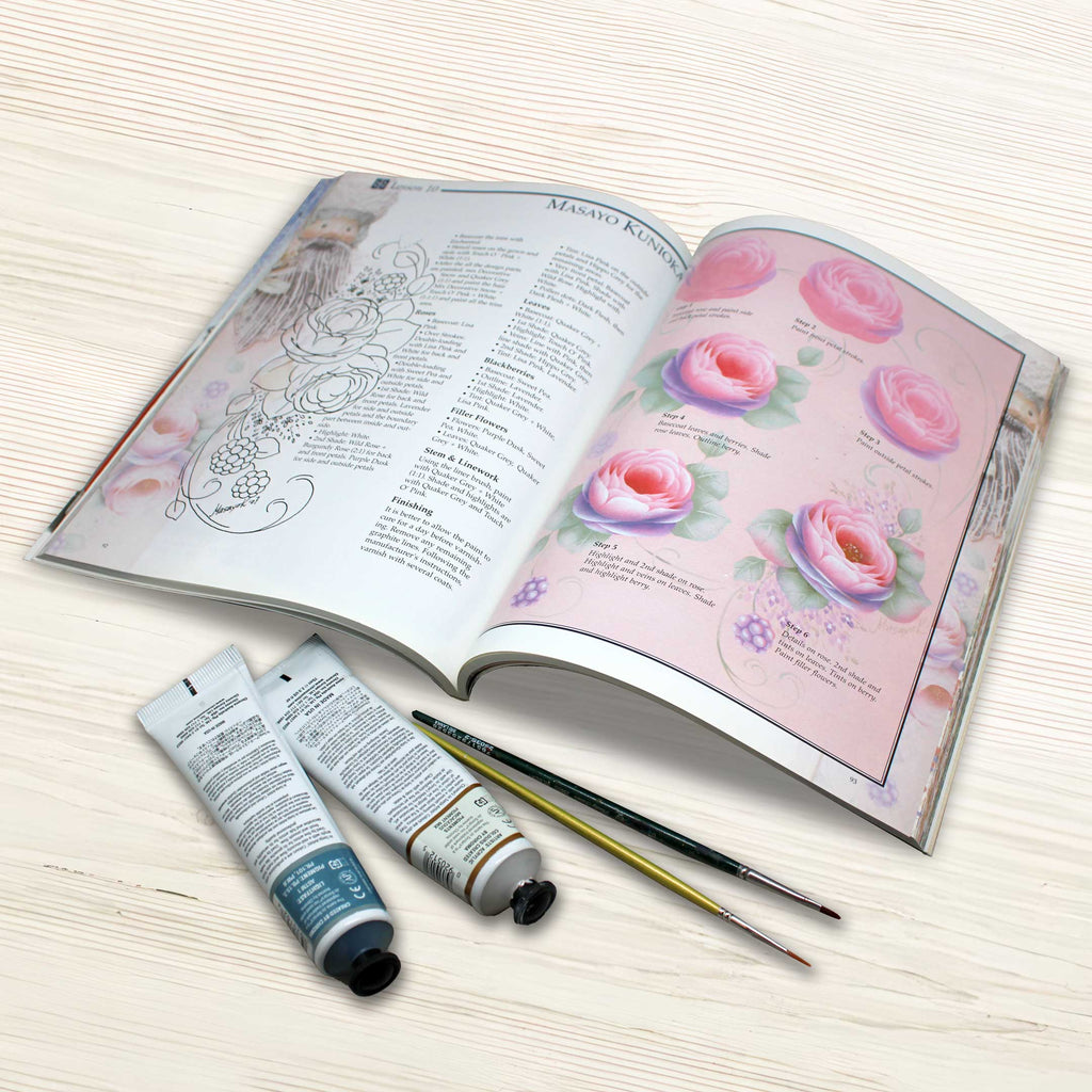 An open book with illustrations of flowers and a list of steps for painting flowers, along with two tubes of paint and two paintbrushes on a wooden surface.