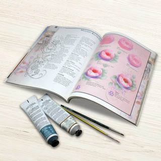 An open book with illustrations of flowers and a list of steps for painting flowers, along with two tubes of paint and two paintbrushes on a wooden surface.